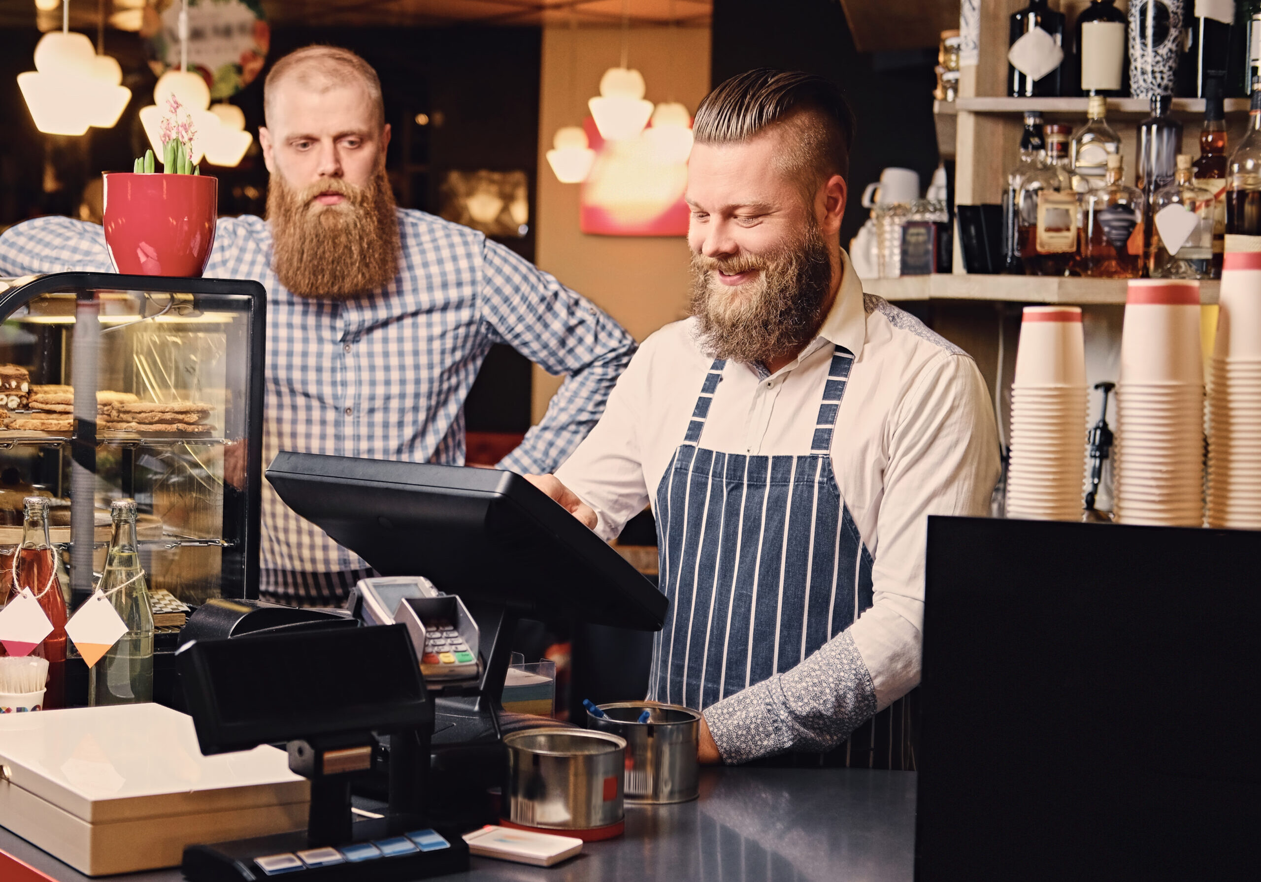 Positive bearded male at the counter using cash register in a coffee shop.